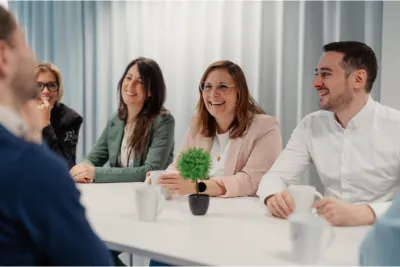 "Groupe de collègues souriants assis autour d'une table blanche, partageant un moment convivial avec des tasses de café, dans un environnement de bureau lumineux