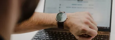 Employee checking a wristwatch while working on a laptop, illustrating Protime time and attendance tracking and time registration