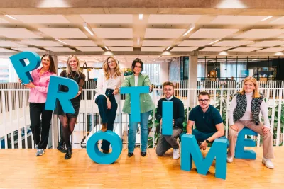 Groep mensen in een kantoor met grote blauwe letters 'PROTIME' in de hand en lachend naar de camera.